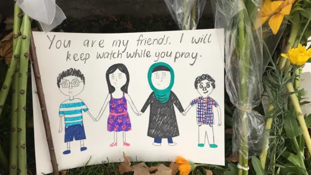 A child's drawing of people holding hands, one in a hijab. says 'you are my friends, I will keep watch while you pray'