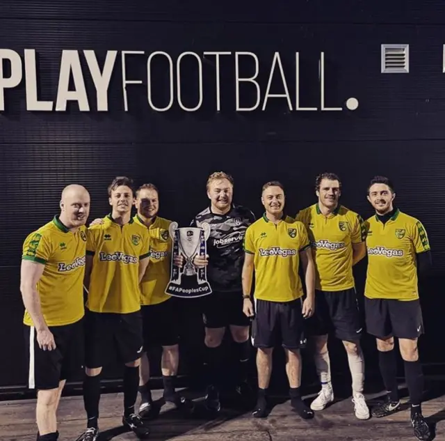 Team photo of an adult male FA People's Cup team in their yellow kit holding a cardboard FA Cup