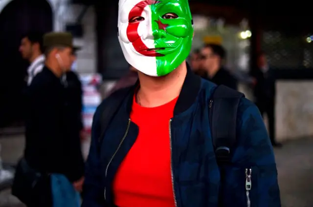 A woman wears a plastic mask printed with the Algerian national flag.