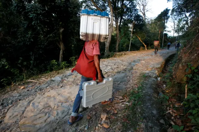 A porter carries a Voter Verifiable Paper Audit Trail (VVPAT) machine and an Electronic Voting Machine (EVM) through Buxa Tiger Reserve forest to a remote polling station, in Alipurduar district in the eastern state of West Bengal