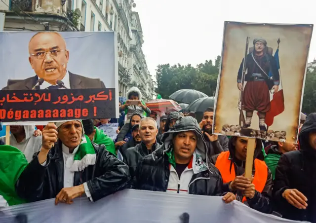 Algerians hold up signs depicting former prime minister Ahmed Ouyahia (R) dressed as a Zouave alongside another showing current PM Noureddine Bedoui with a caption below reading in Arabic "biggest election forger", during an anti-government demonstration in the northeastern city of Annaba on 12 April 2019.