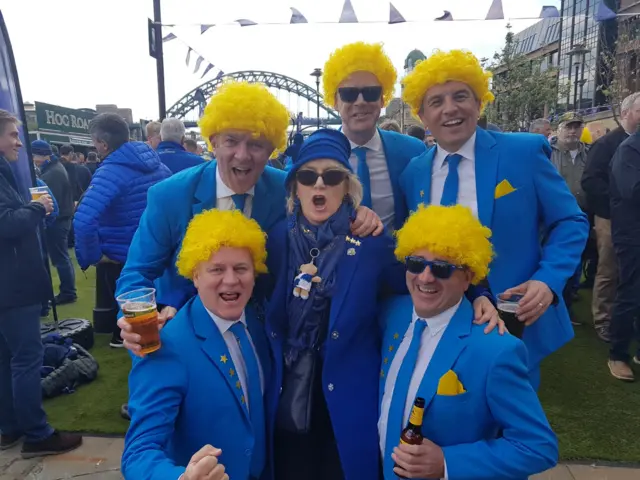 Fans dressed in blue suits with yellow wigs and a fan in the middle in a blue coat and blue hat