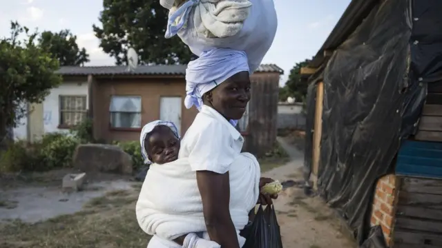 A smiling woman carrying a baby on her back and a bag on her head