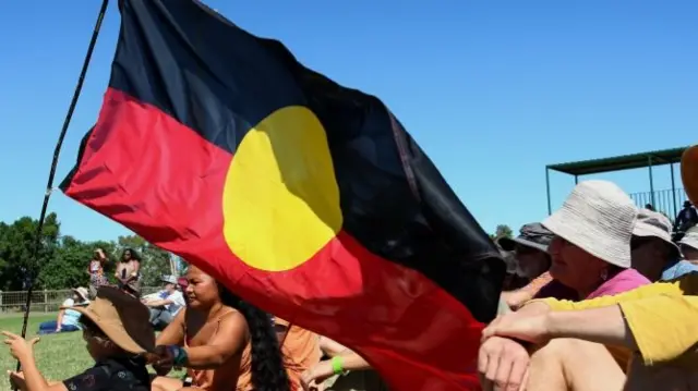 A young boy sitting in crowd holds up the Aboriginal flag