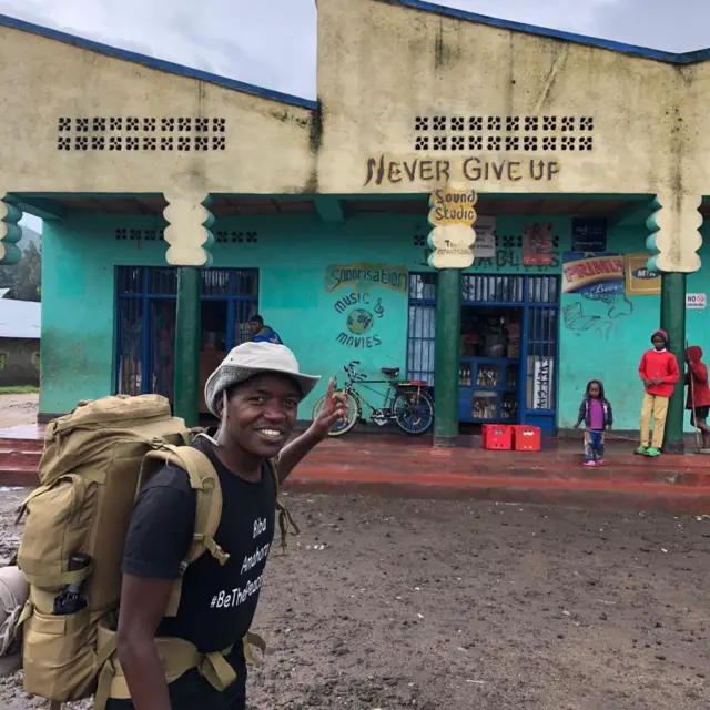 Hyppolite Ntigurirwa pointing to type on a building wthat reads 'Never Give Up'.