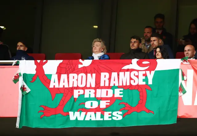 Aaron Ramsey sitting in the stands at the Emirates Stadium behind a banner that says: "Aaron Ramsey - pride of Wales."