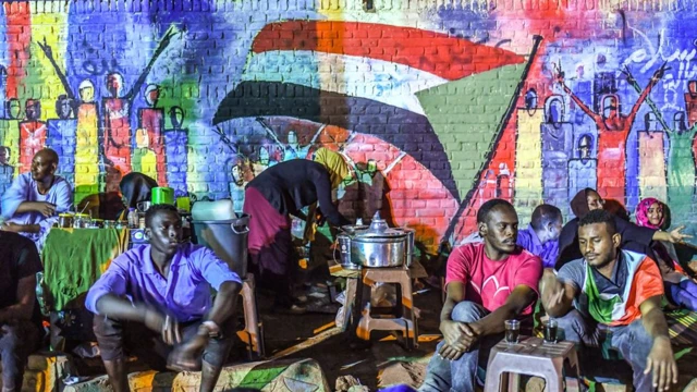 Protesters in Sudan pictured at night by the military headquarters, sitting in front of a mural