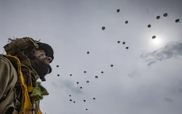 Dakota planes drop parachutists in Second World War attire along the Normandy coast.