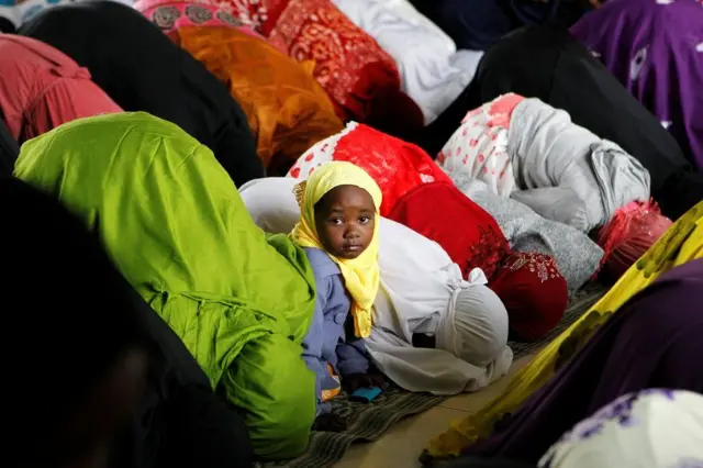 Girl looking at the camera whilst women around her are praying