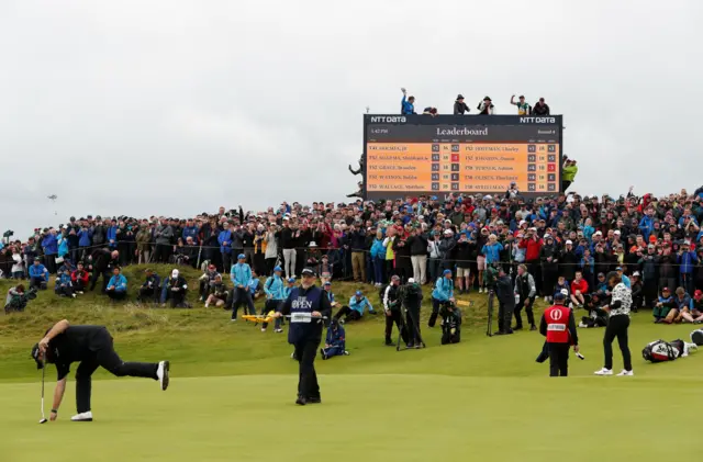 Fans are partying on the scoreboard as Shane Lowry bends to retrieve his ball from the hole on the 16th