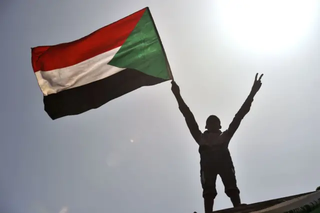 An anonymous protester standing on the roof of a building flies the Sudanese flag