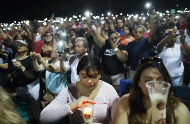 People hold up their phones in lieu of candles at an interfaith vigil