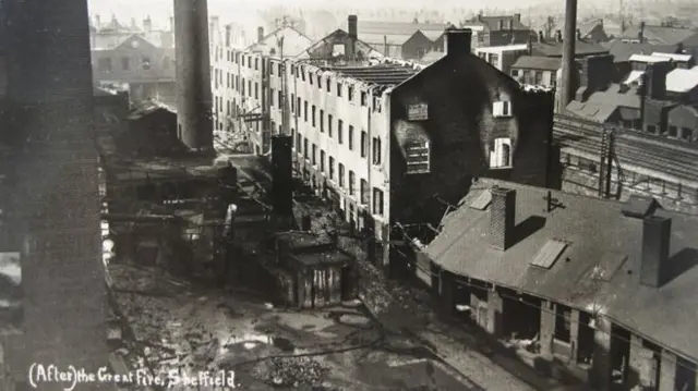 A black and white postcard showing the great fire of Heeley in 1921. A burnt out building is shown with a heavily damaged roof. A chimney stack is to the left of the building with more damage being shown. The Midland Mainline railway is to the right of the building, which is still in use today, that took passengers from Sheffield to London.