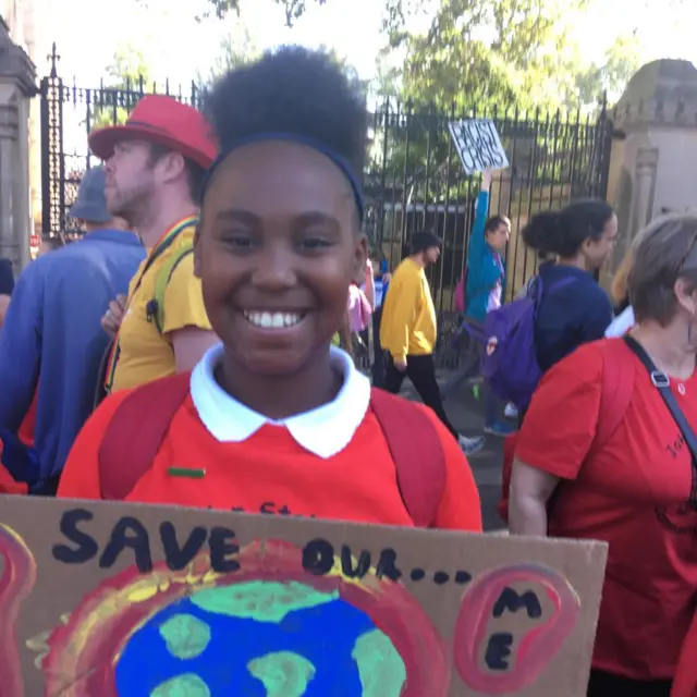 Mariah wears a red school uniform and holds a placard