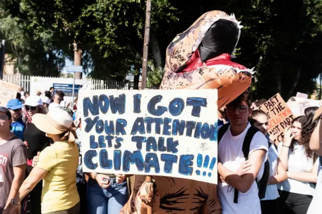 A person dressed as a dinosaur holding a sign that says: "Now I got your attention let's talk about climate!!!"