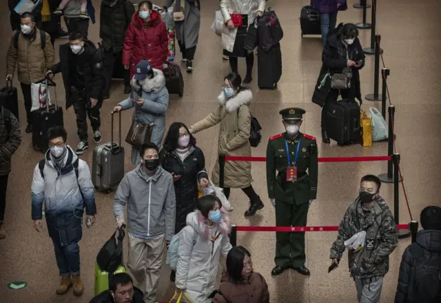 A police officer in ornate uniform wears a cheap filter mask, standing to attention amid a crowded train station