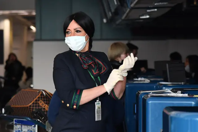 A female airport employee in uniform, and masked, pulls on surgical gloves to inspect baggage