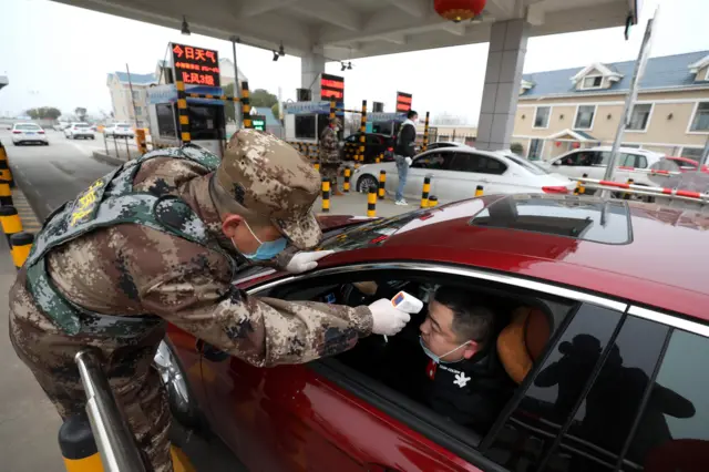 A man sitting in a car has his temperature taken by a uniformed soldier in front of a checkpoint