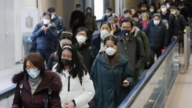 Dozens of passengers at an airport are all seen wearing face masks in a queue stretching into the distance