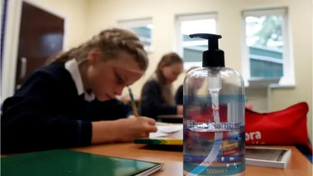 A school pupil writers at a desk next to a bottle of hand sanitiser