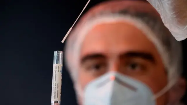 A health worker, wearing a protective suit and a face mask, holds a test tube after administering a nasal swab to a patient