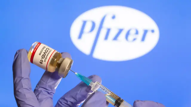 A woman holds a small bottle labeled with a "Coronavirus COVID-19 Vaccine" sticker and a medical syringe in front of displayed Pfizer logo in this illustration taken, October 30, 2020.