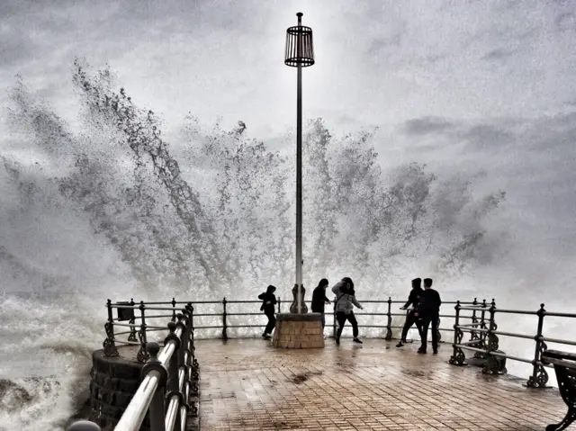 Wave breaks over sea wall and people