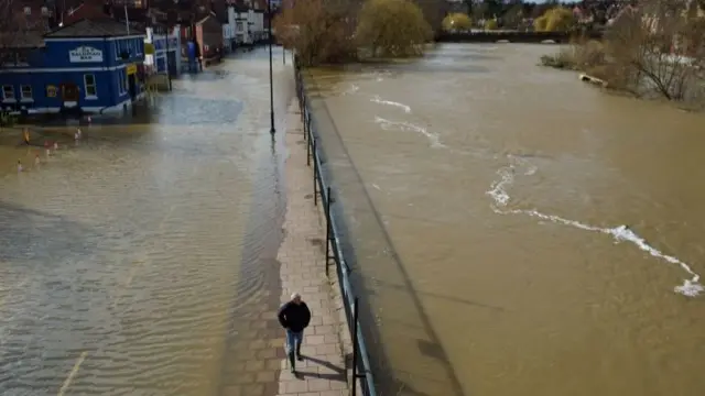 A man makes his way carefully between the flooded Smithfield Road in Shrewsbury (left) and the River Severn (right),