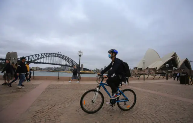 A cyclist with a face mask in front of the Sydney Opera House and Sydney Harbour Bridge