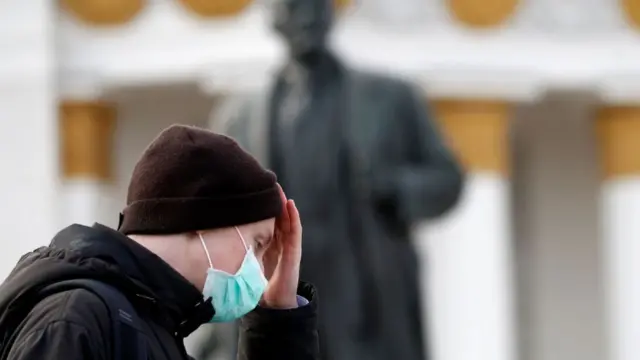 A young man wearing a medical masks walks past a statue of Lenin in Moscow