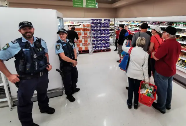 Police officers stand next to a queue of shoppers waiting to buy toilet paper