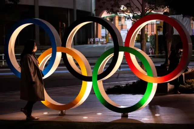 A woman wearing a protective face mask walks past The Olympic rings in front of the Japan Olympics Museum in Tokyo, Japan, March 3, 2020