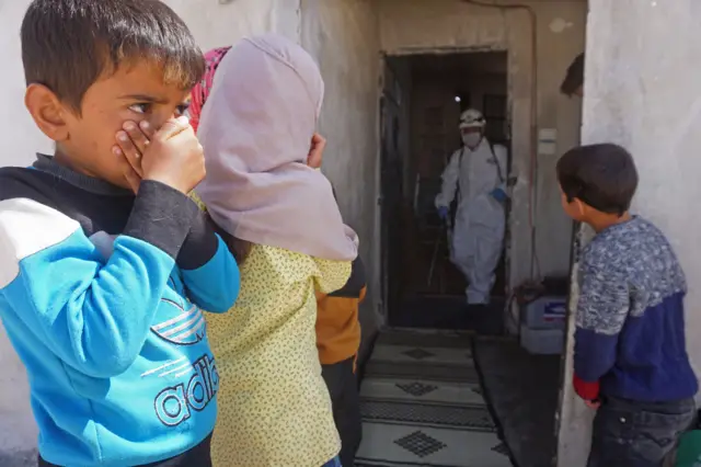 Children cover their faces as White Helmet health workers disinfect a building in Binnish in rebel-held Idlib (26/03/20)