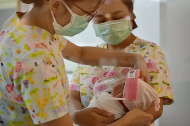 This photo taken through a glass window at a maternity ward shows nurses holding newborn babies wearing face shields