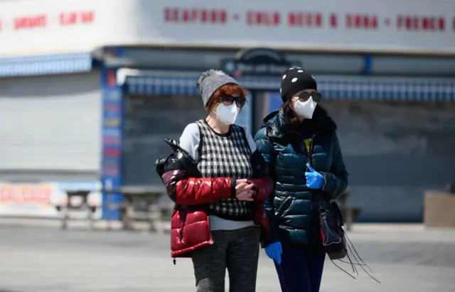 Two women in face masks walk down the Coney Island boardwalk