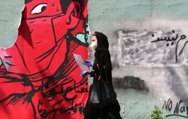 A Iranian woman wearing a protective mask and gloves walks past a mask graffiti in Tehran