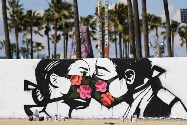 Palm trees stand behind a mural depicting two people kissing while wearing face masks on a beach in California