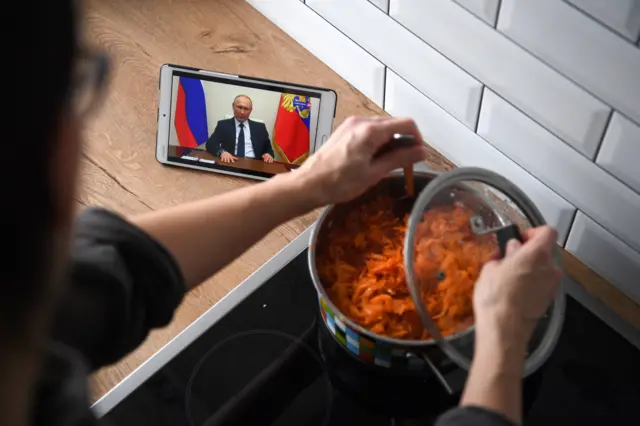 A woman in Russia cooking while watching President Putin give a speech on her phone