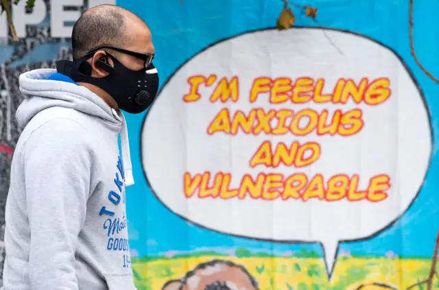 A man wearing a face mask past a poster saying 'I'm Feeling Anxious and Vulnerable'