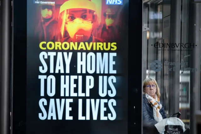 A woman waits at a bus stop on Edinburgh's Princes Street next to a coronavirus advert