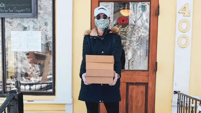 A woman wearing a face mask and large coat holds four boxes of pies outside a house