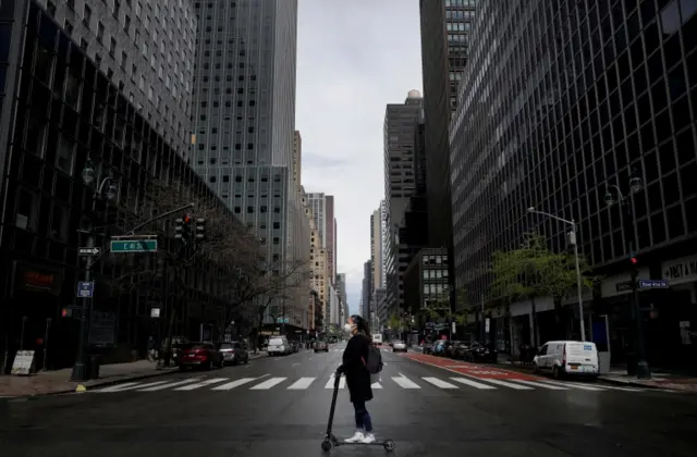 A woman wearing a protective face mask rides a scooter across a nearly empty 3rd Avenue in midtown Manhattan