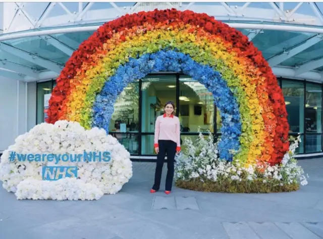 Woman standing under a rainbow made of flowers. A white flower cloud reads: #weareyourNHS