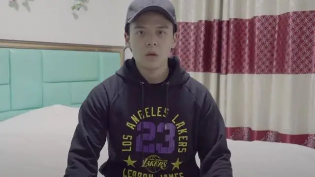 Screenshot of Li Zehua shows him speaking to camera, wearing a hoodie and a cap