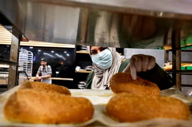 A Palestinian woman, wearing protective masks due to the COVID-19 coronavirus pandemic, picks out freshly-baked pastries from a tray as she shops for groceries in the city of Hebron in the occupied West Bank on April 23, 2020