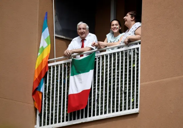 A family in Milan stand on their balcony behind an Italian flag