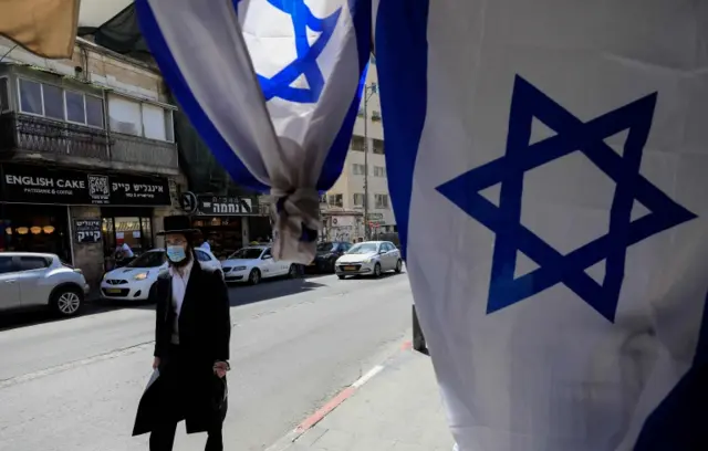 A man wearing a mask walks past an Israel flag