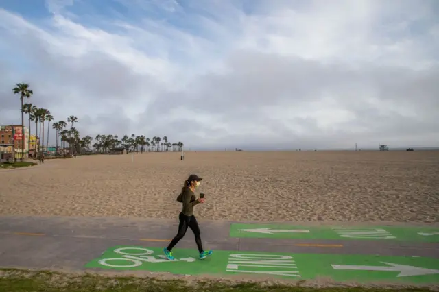 A woman wearing a mask, runs on the bike path along the deserted beach in Venice, California