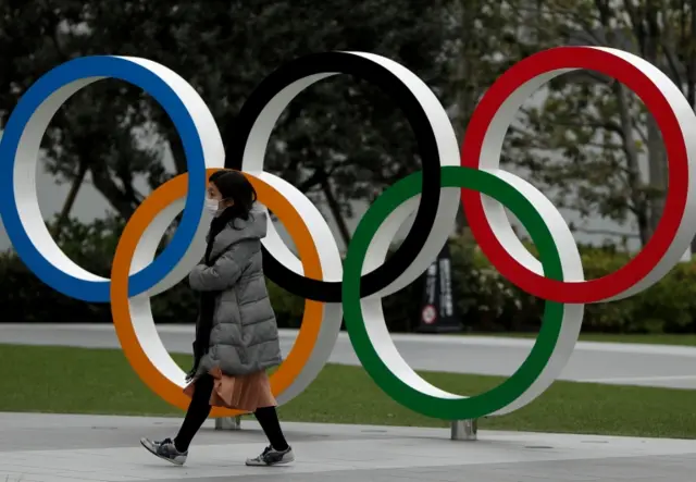 A woman wearing a mask walks past the Olympic rings