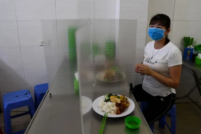 A woman, wearing a face mask eats lunch on a table installed with a make-shift plastic partition, Hanoi